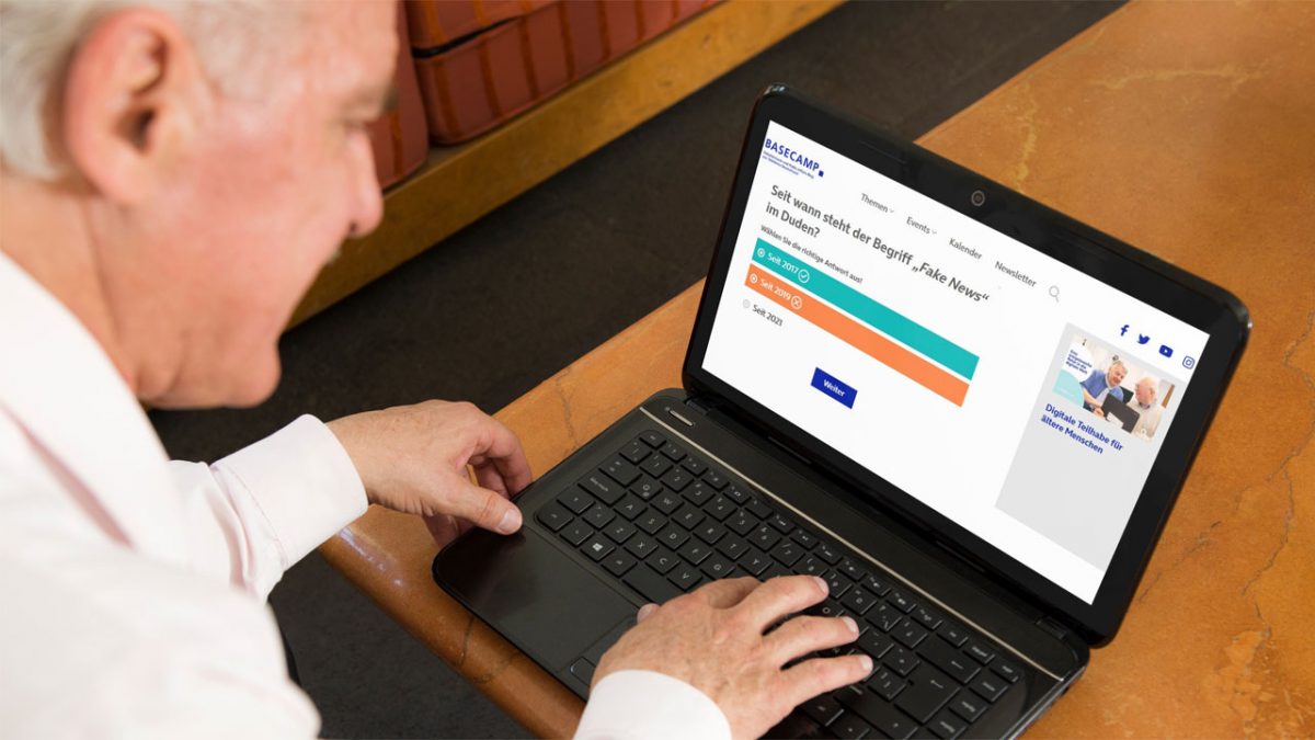 Älterer Mann sitzt am geöffneten Laptop am Schreibtisch. Geöffnet ist die Seite vom Basecamp Quiz "Fit gegen Fake News" und der Frage "Seit wann steht der Begriff "Fake News" im Duden?